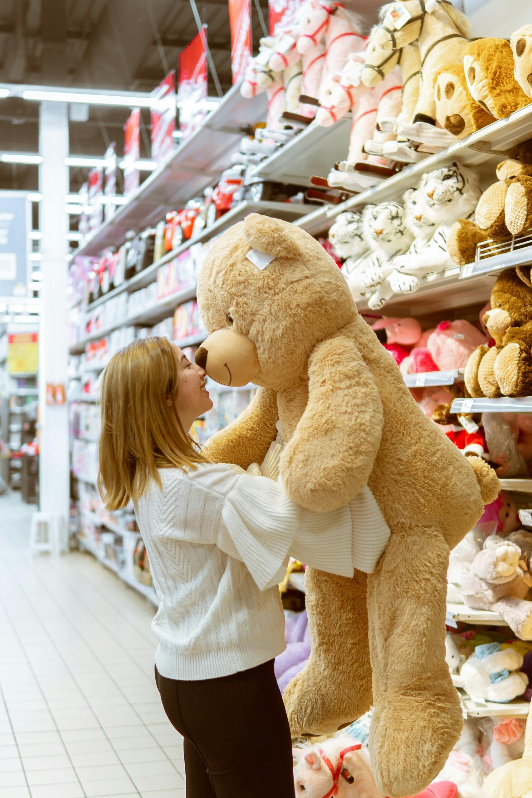 A woman chooses a large teddy bear in a retail toy section, surrounded by stuffed animals.